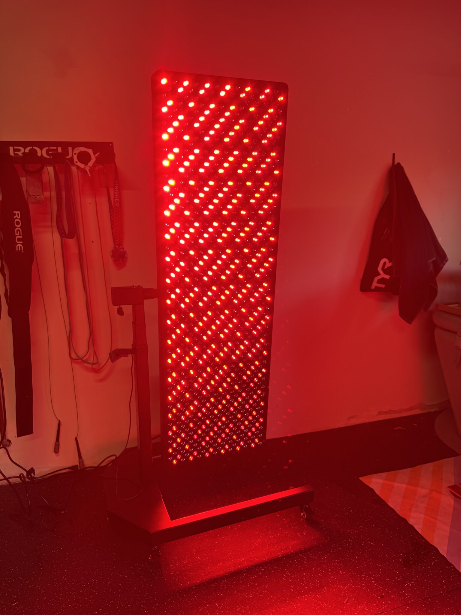 A large standing red light therapy panel is illuminated in a room, casting a red glow on the surrounding area; workout gear hangs on the wall nearby.
