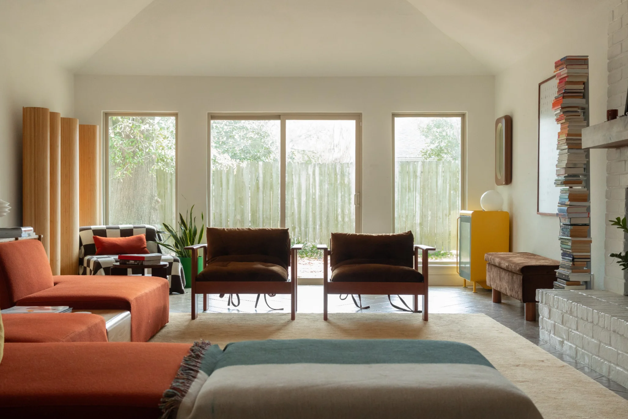A living room with modern furniture, large windows overlooking a fenced yard, a stack of books on a white fireplace, and several plants.