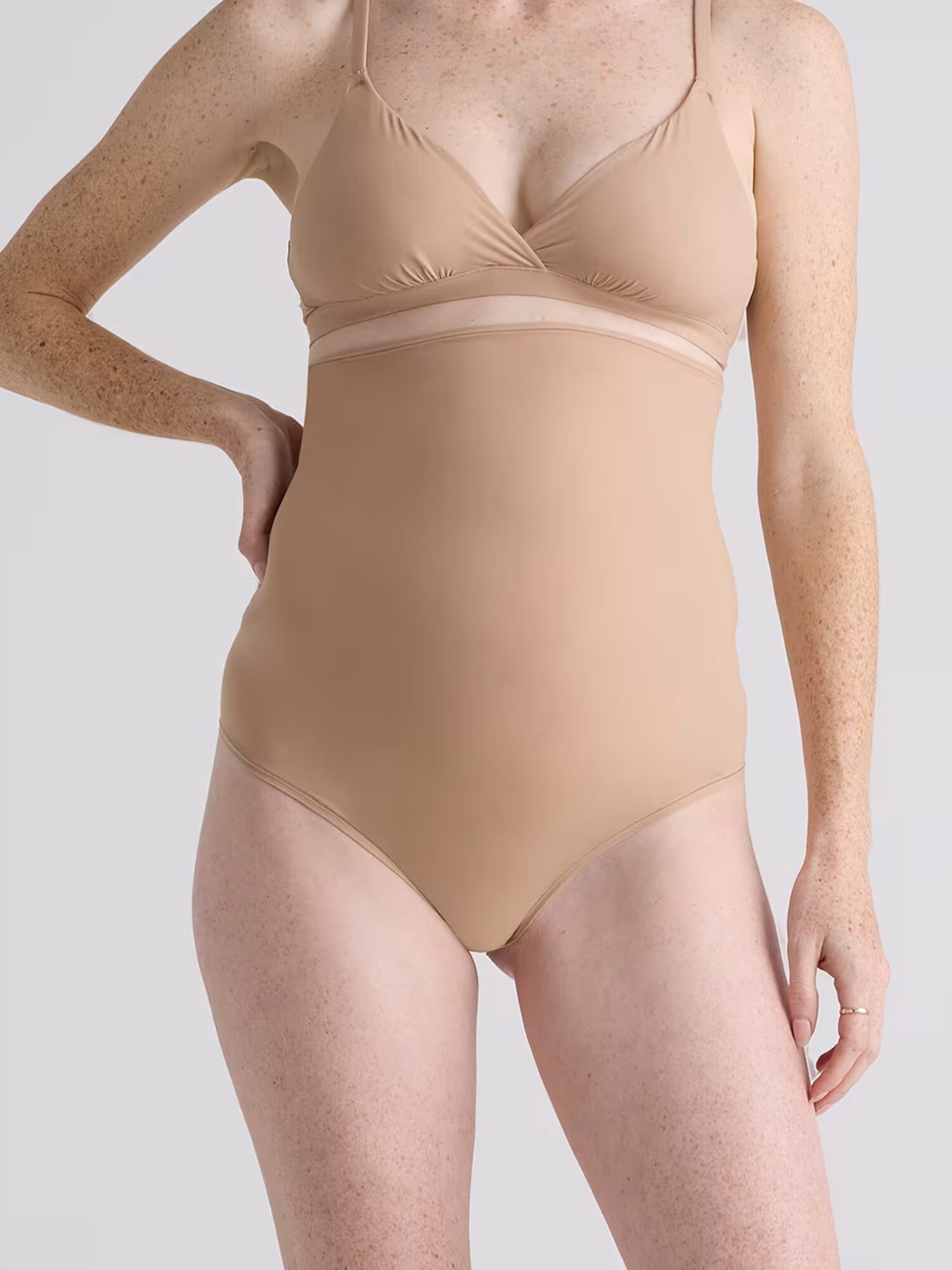 A person wearing a nude-colored, seamless one-piece bodysuit stands against a plain light gray background.