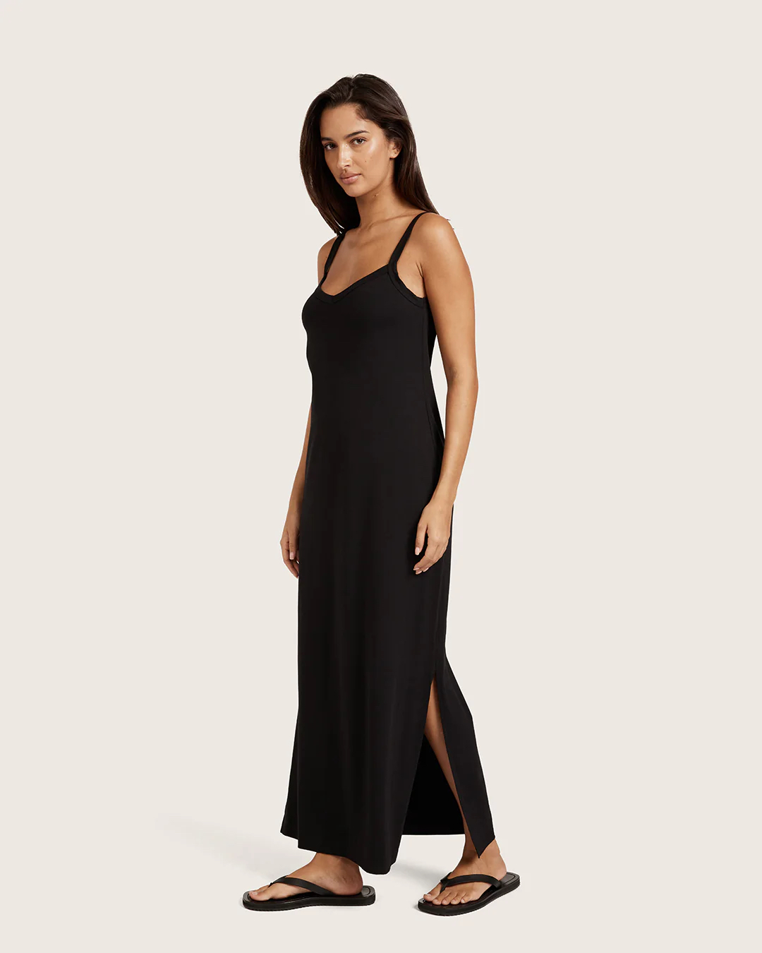 Woman standing and wearing a black sleeveless maxi dress with thin straps and side slit, paired with black sandals, against a plain light background.