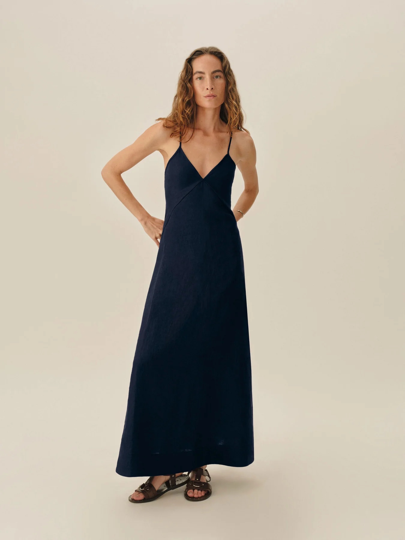 Woman standing in a navy blue sleeveless maxi dress with thin straps and sandals, against a plain light background.
