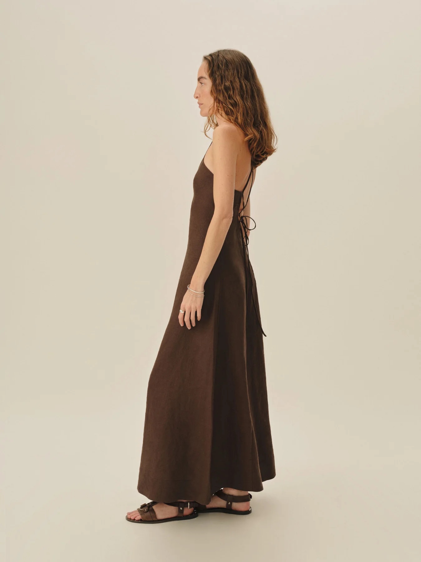 Woman standing sideways wearing a sleeveless, back-tied, brown maxi dress and black sandals against a plain beige background.