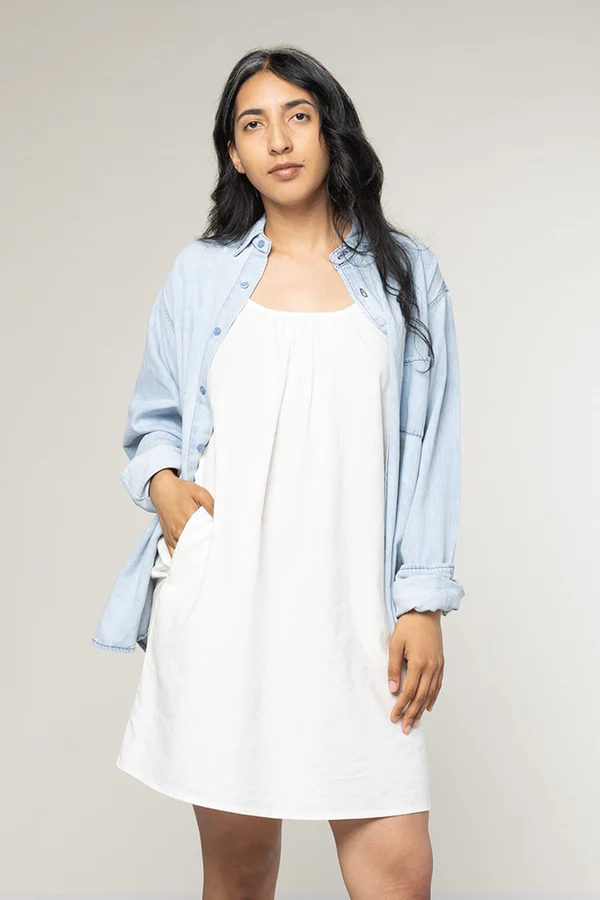 Person standing against a plain background, wearing a white dress with a light blue button-up shirt over it, one hand in dress pocket.