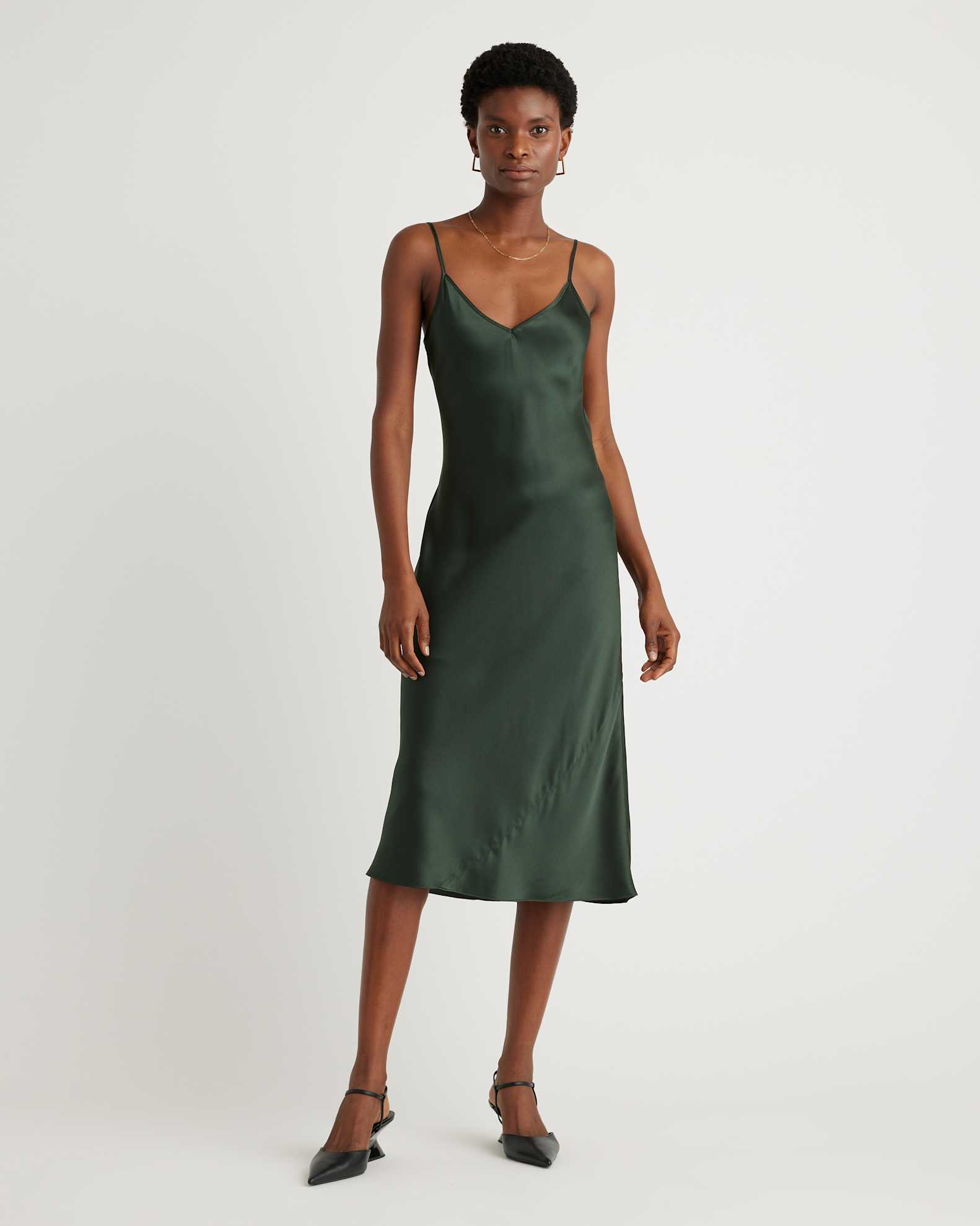 A woman stands against a plain white background wearing a dark green satin slip dress with thin straps and black heels.