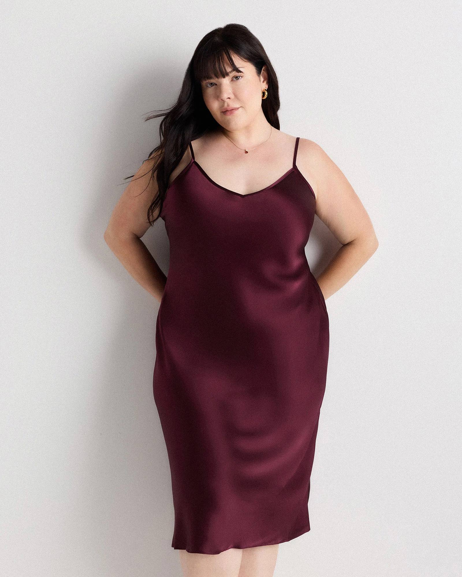 Woman with long dark hair stands against a plain light wall, wearing a sleeveless burgundy satin slip dress and a small necklace, looking directly at the camera with a neutral expression.