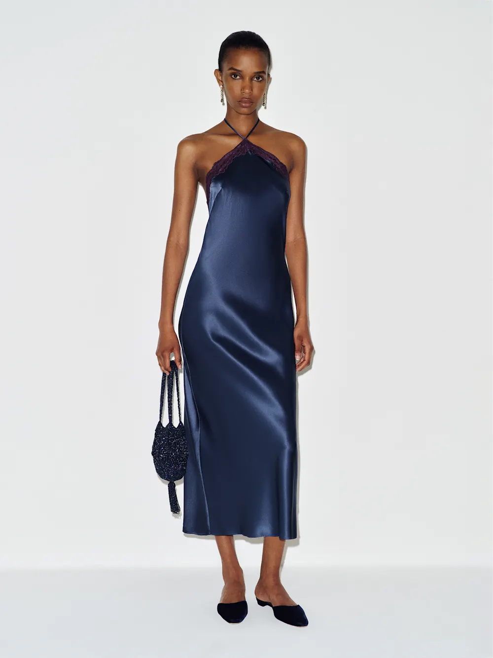 A woman stands against a white background wearing a navy satin halter dress, black flat shoes, and holding a dark woven handbag.