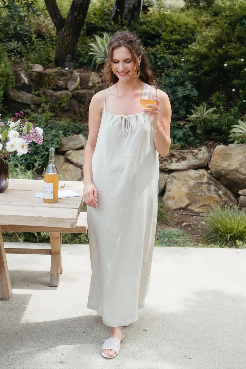A woman in a light linen dress stands outdoors holding a drink, next to a wooden table with a bottle and flowers, with greenery and rocks in the background.