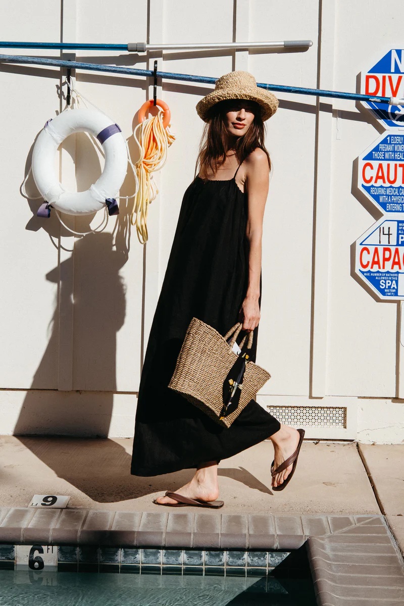 A person in a black maxi dress, flip-flops, and straw hat stands by a pool holding a woven bag. Safety signs and pool equipment are visible in the background.