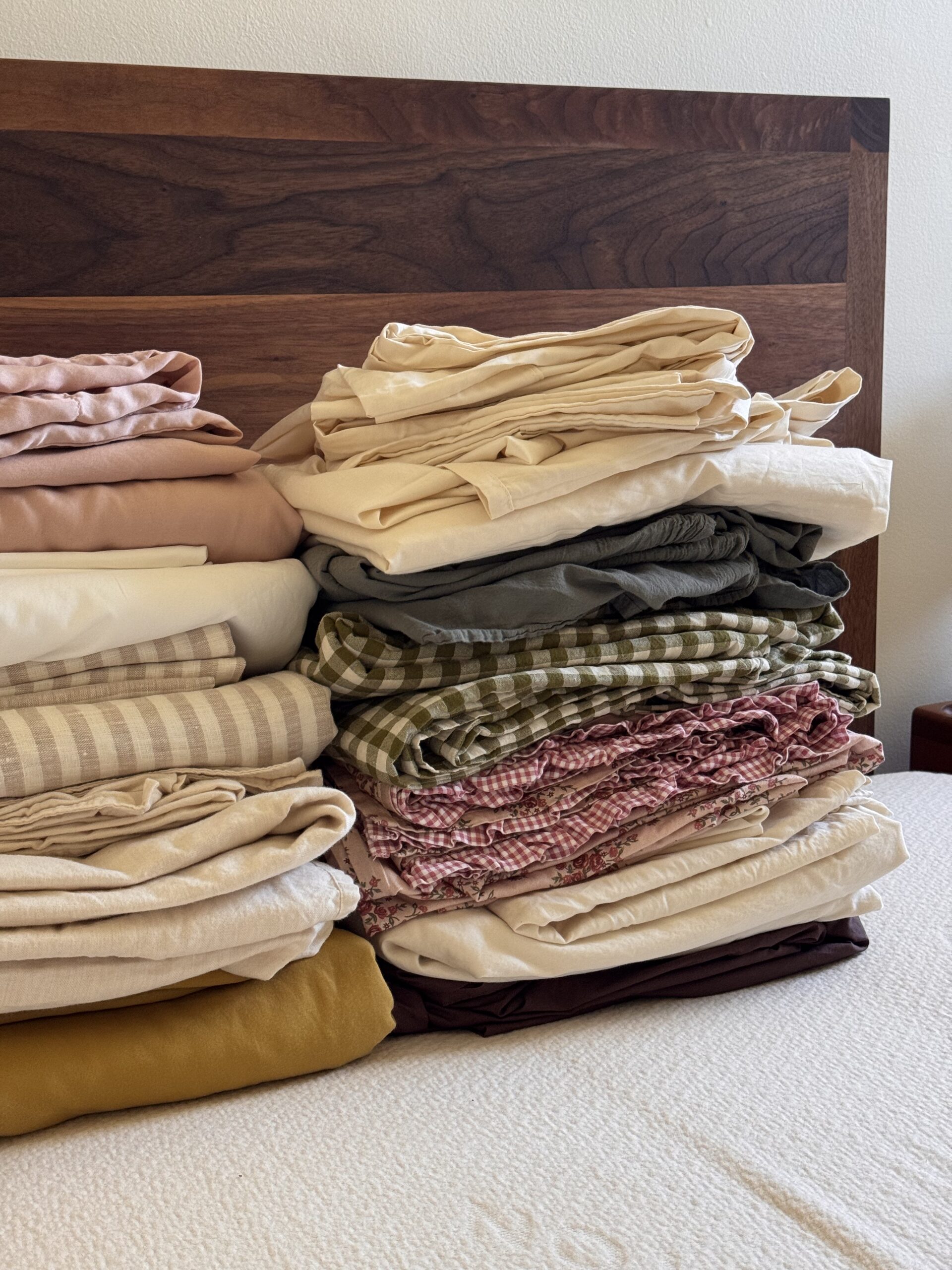 Neatly folded stacks of bed sheets and pillowcases in various colors and patterns are arranged on a bed in front of a wooden headboard.