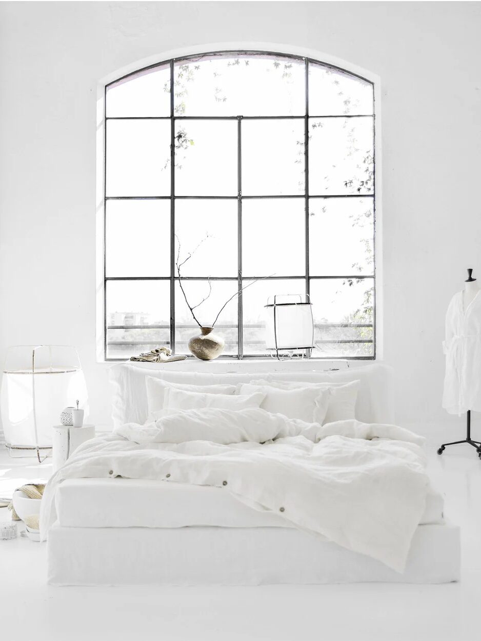 A minimalist white bedroom with an unmade bed, large industrial-style window, simple decor, and a dress form with a robe.