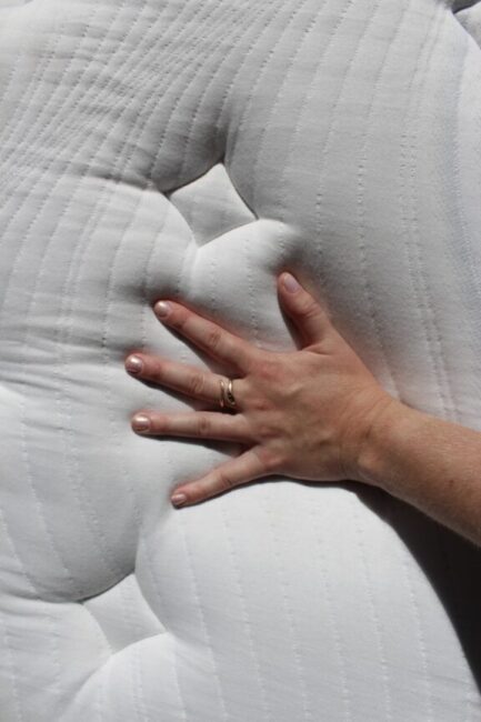 A hand with a gold ring presses into a white, quilted mattress, creating an indentation on the fabric surface.
