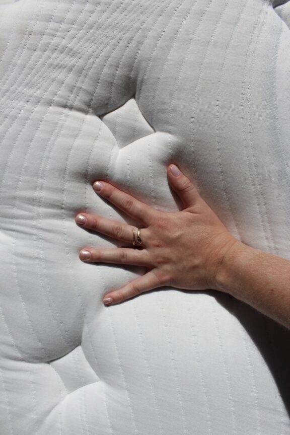 A hand with a gold ring presses into a white, quilted mattress, creating an indentation on the fabric surface.