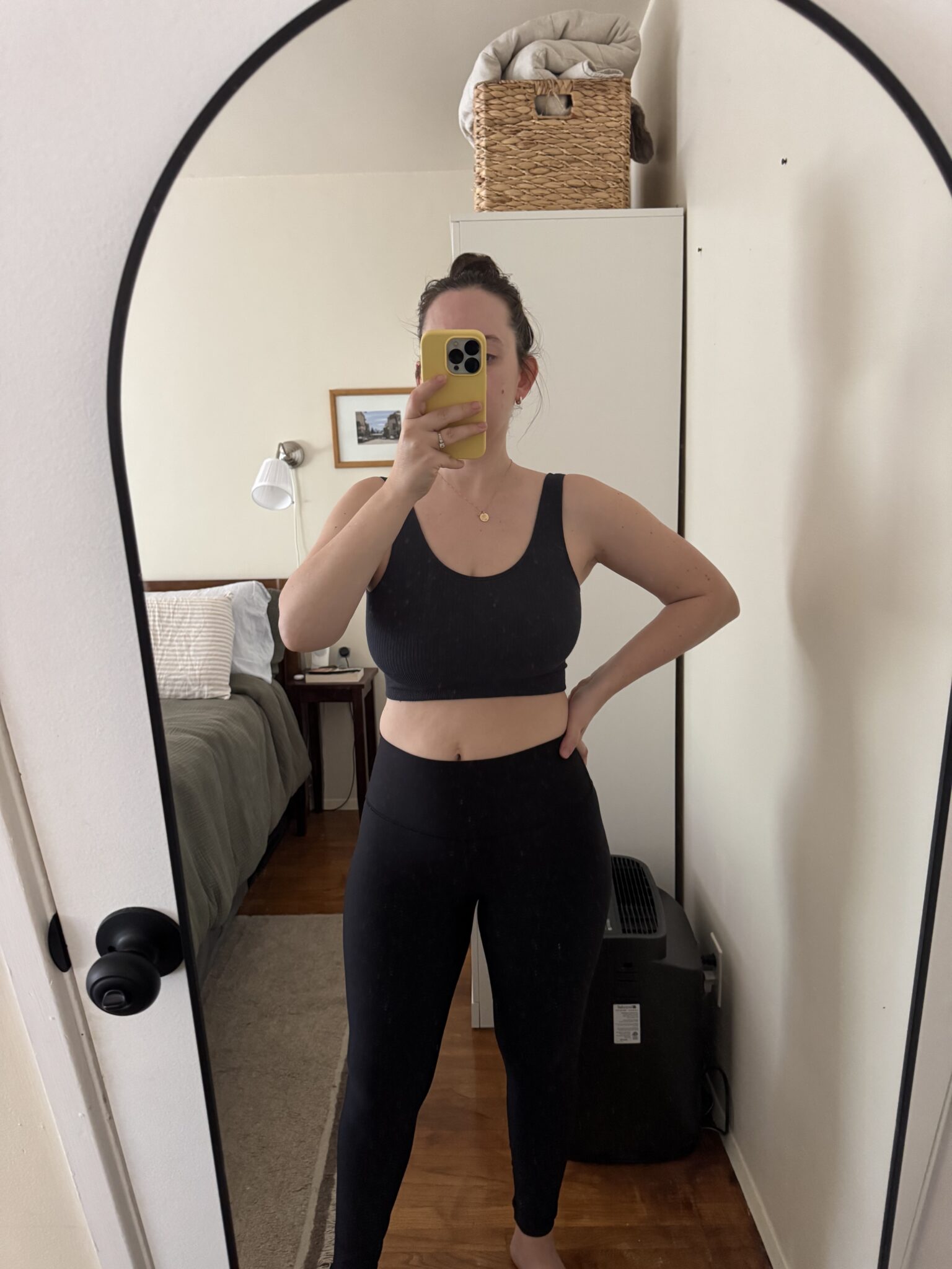 A person wearing a black sports bra and leggings takes a mirror selfie in a bedroom with a bed, lamp, and basket in the background.