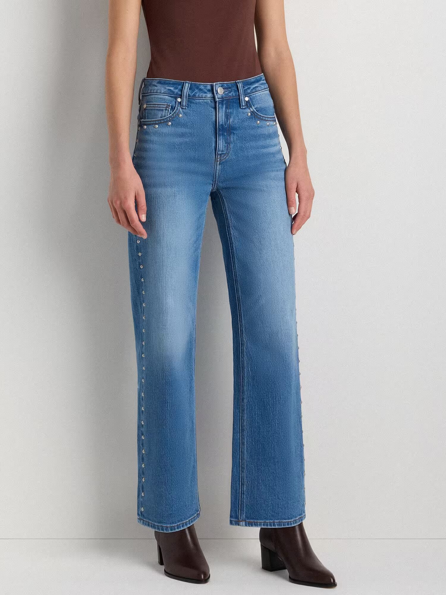 A person wearing light blue wide-leg jeans with rivet details on the side seams, a brown top, and brown ankle boots, standing against a plain light wall.