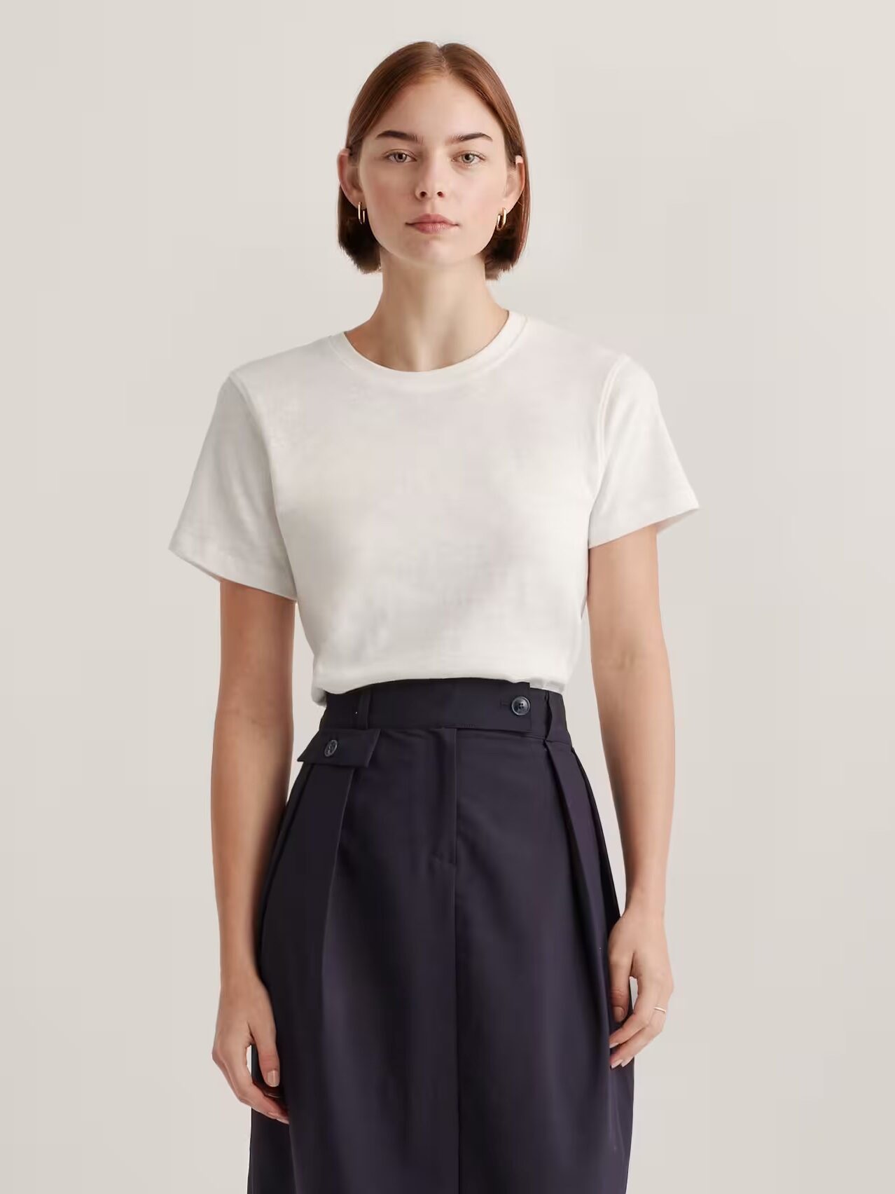 A person stands against a plain background, wearing a white short-sleeve t-shirt tucked into high-waisted, dark navy trousers.