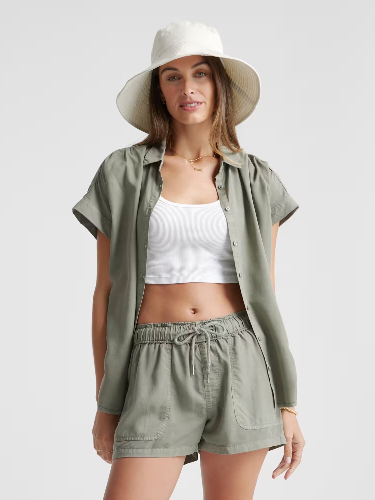 A woman wearing a wide-brim hat, an olive-green short-sleeve button-up shirt, matching drawstring shorts, and a white cropped tank top stands facing forward.