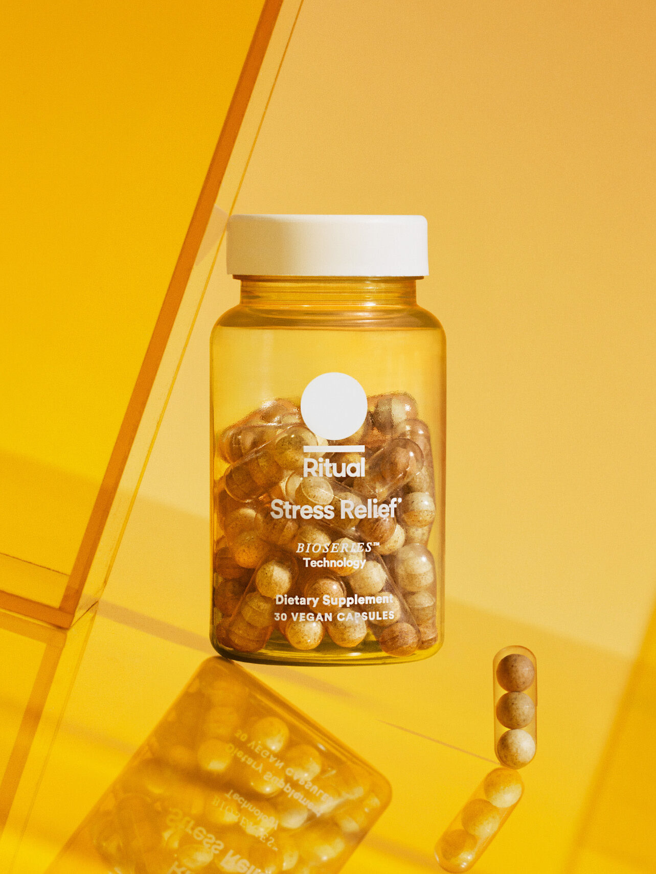 A clear bottle labeled "Ritual Stress Relief" containing brown capsules, placed on a yellow surface with a few capsules outside the bottle.