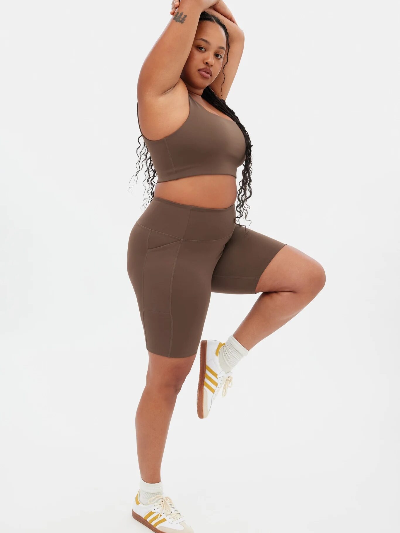 A person wearing a brown sports bra and matching bike shorts balances on one leg with arms raised, against a plain white background.