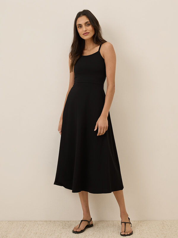 Woman wearing a sleeveless, black, midi-length dress with thin straps and black sandals, standing against a plain light-colored wall.