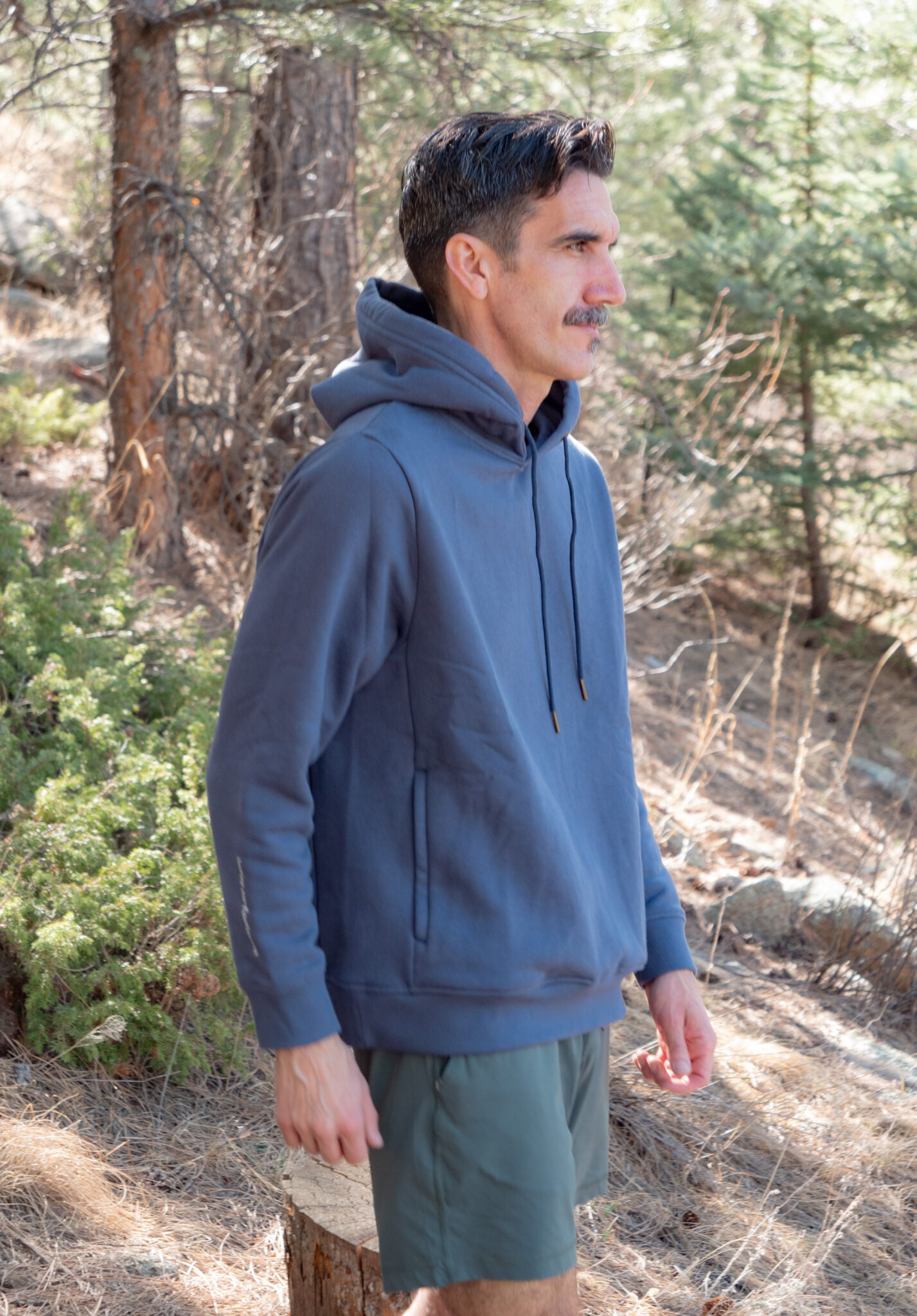 A man wearing a blue hoodie and green shorts stands outdoors in a forested area with trees and dry grass.