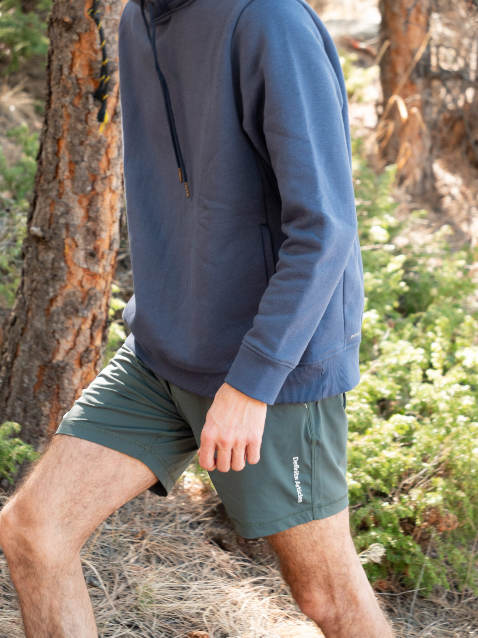 A person wearing a blue hoodie and green shorts is walking outdoors near trees and greenery.