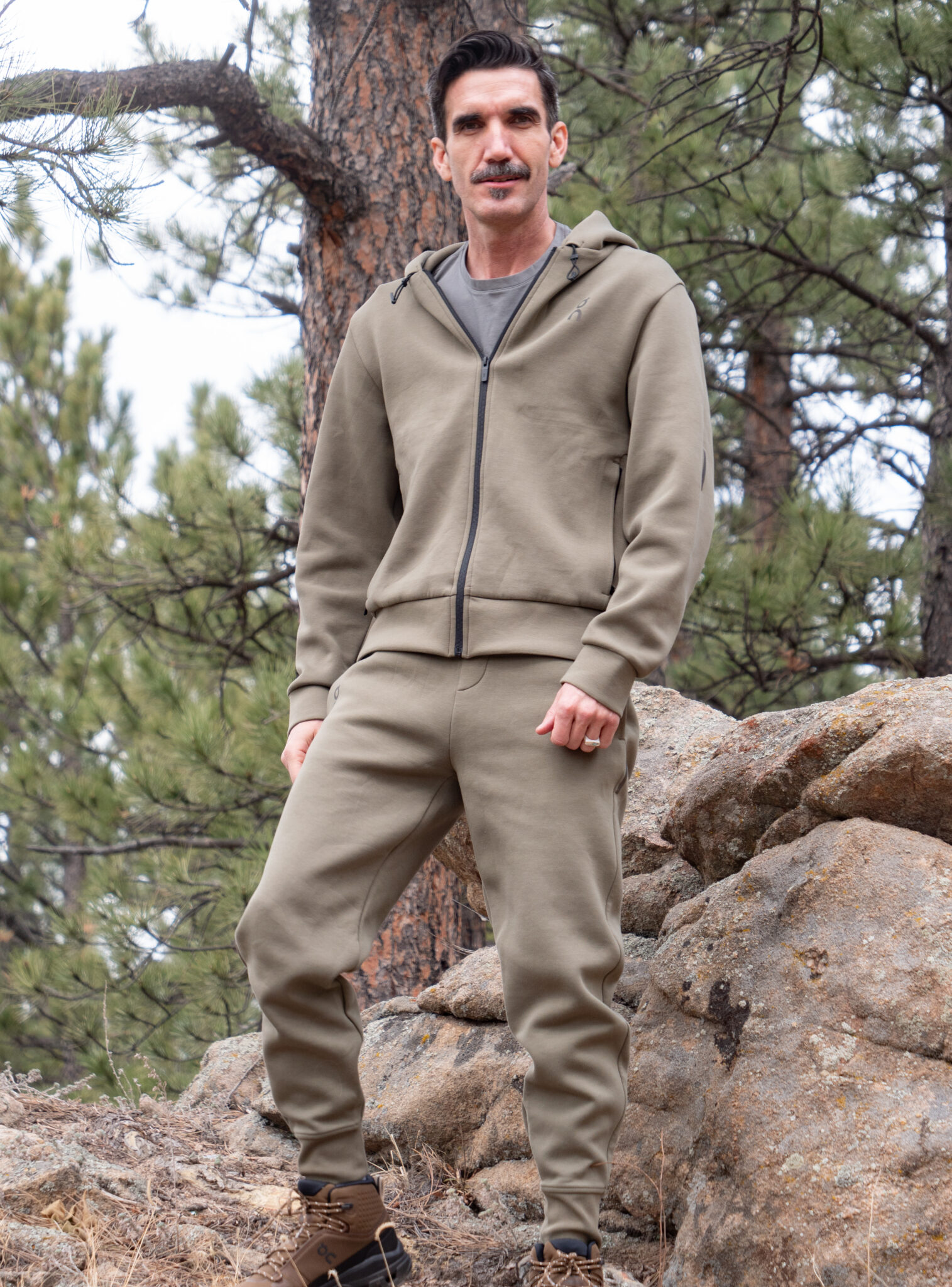 A man wearing an olive green tracksuit and brown boots stands on rocks in a forested area with pine trees in the background.