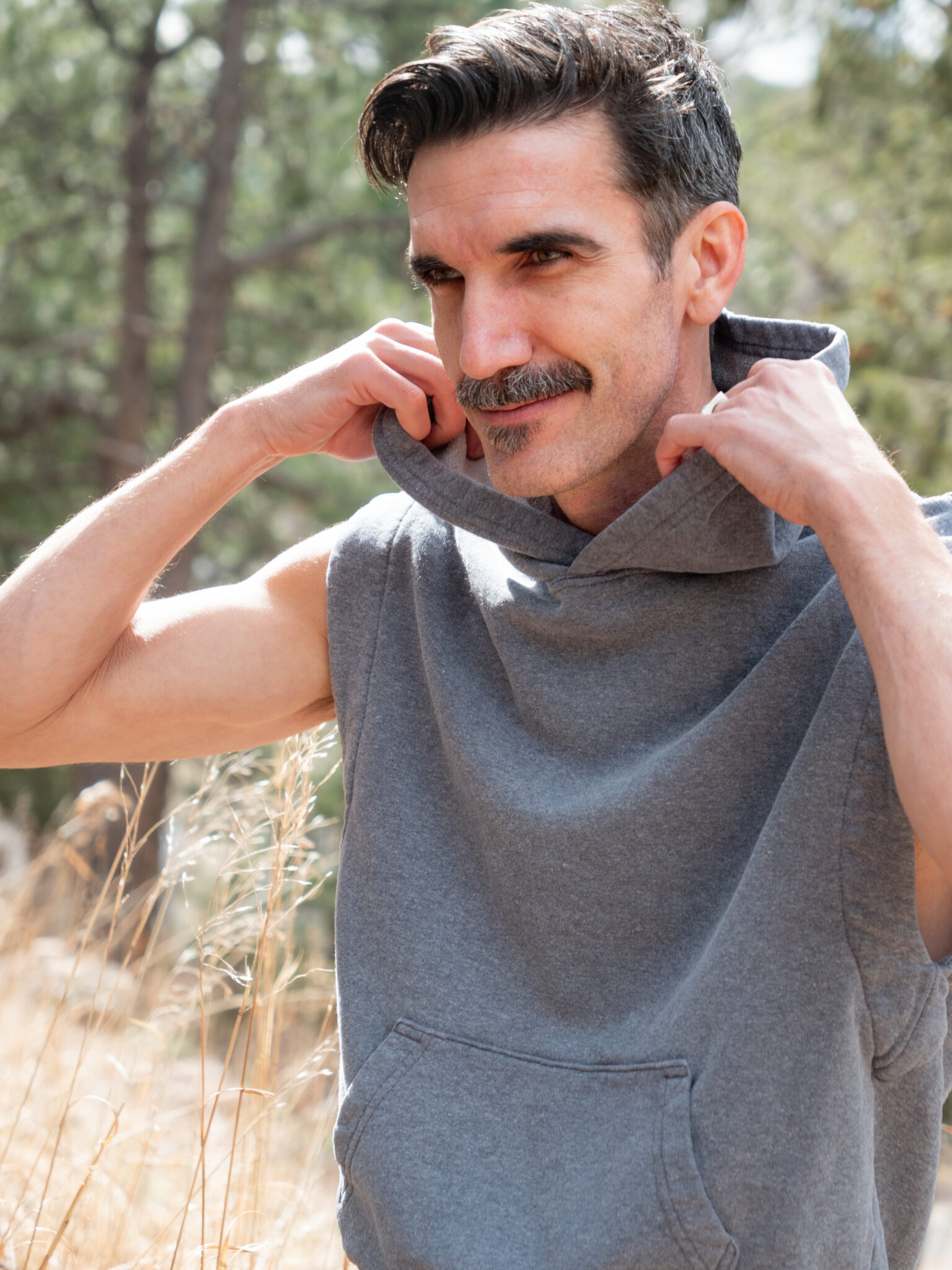 A man with a mustache adjusts the hood of his sleeveless gray hoodie while standing outdoors in a natural setting.