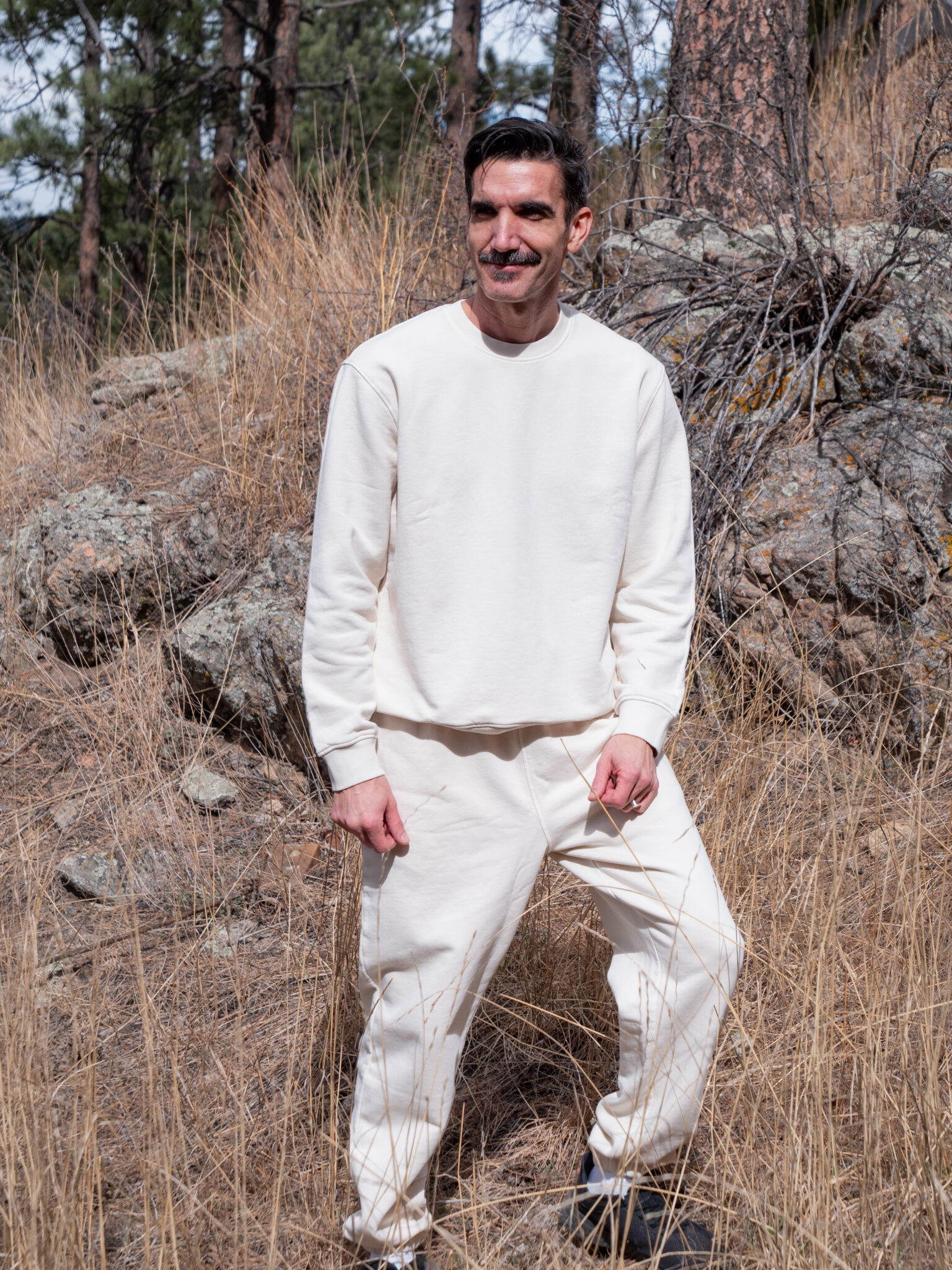 Man standing outdoors in tall dry grass and rocks, wearing a cream-colored sweatshirt and matching pants, with trees in the background.
