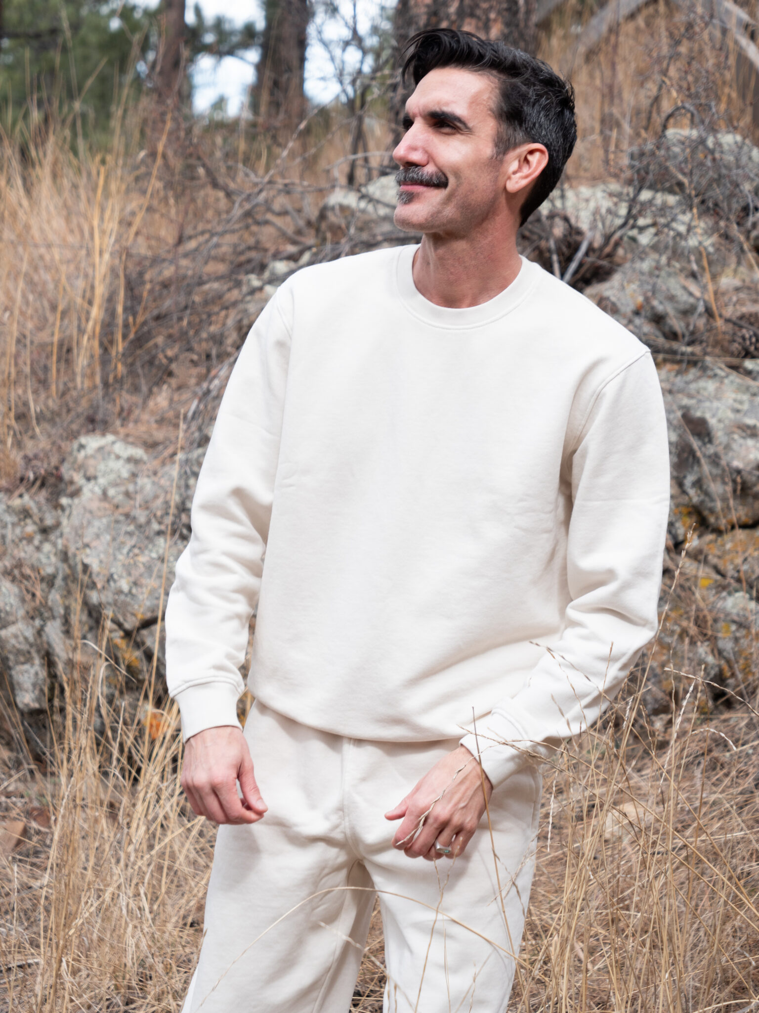 A man wearing an off-white sweatshirt and matching pants stands outdoors among dry grass and rocks, looking to the side and smiling.