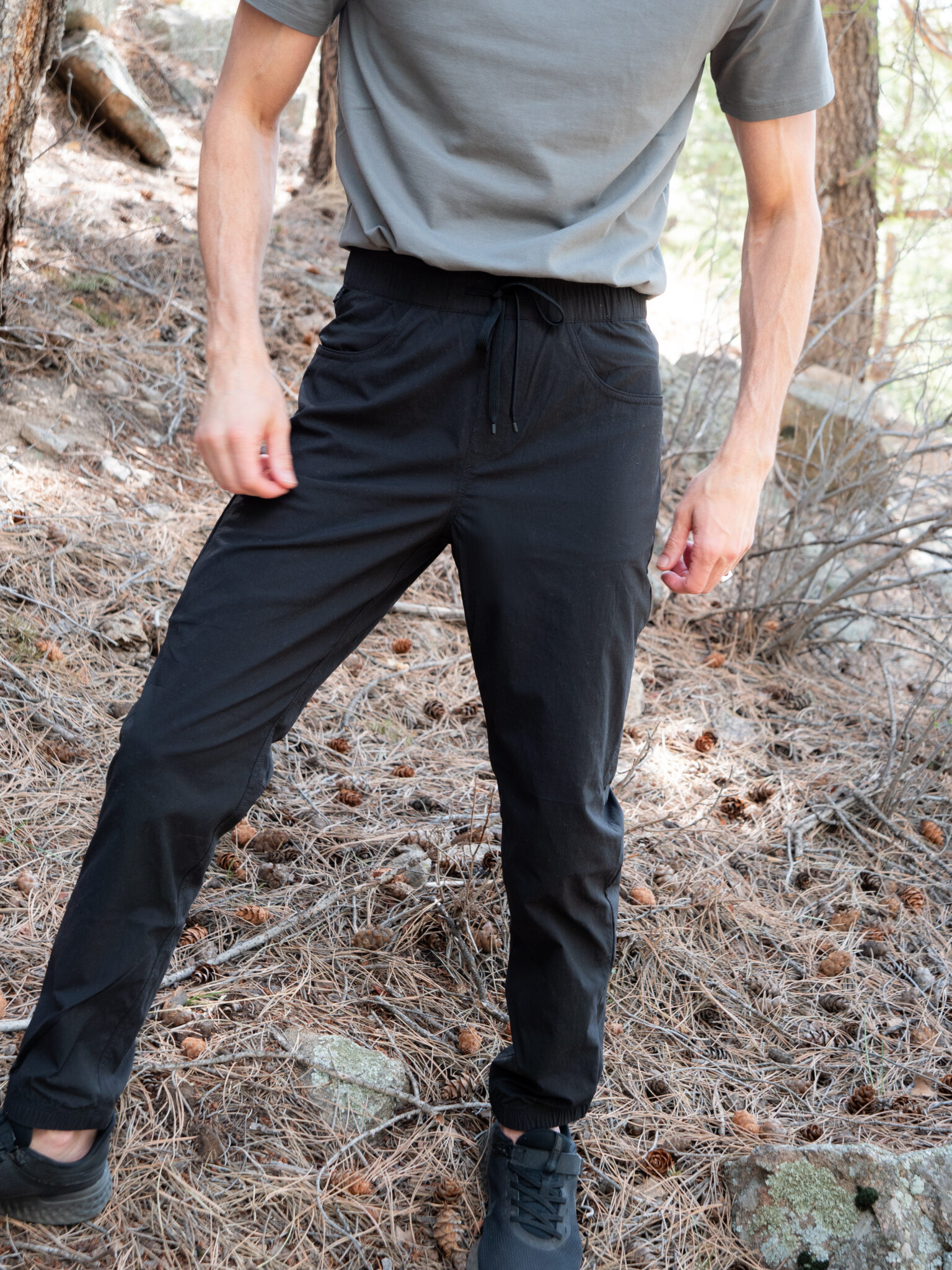 A person standing outdoors on a forest floor, wearing a gray t-shirt, black pants with a drawstring, and black shoes. Their face is not visible.