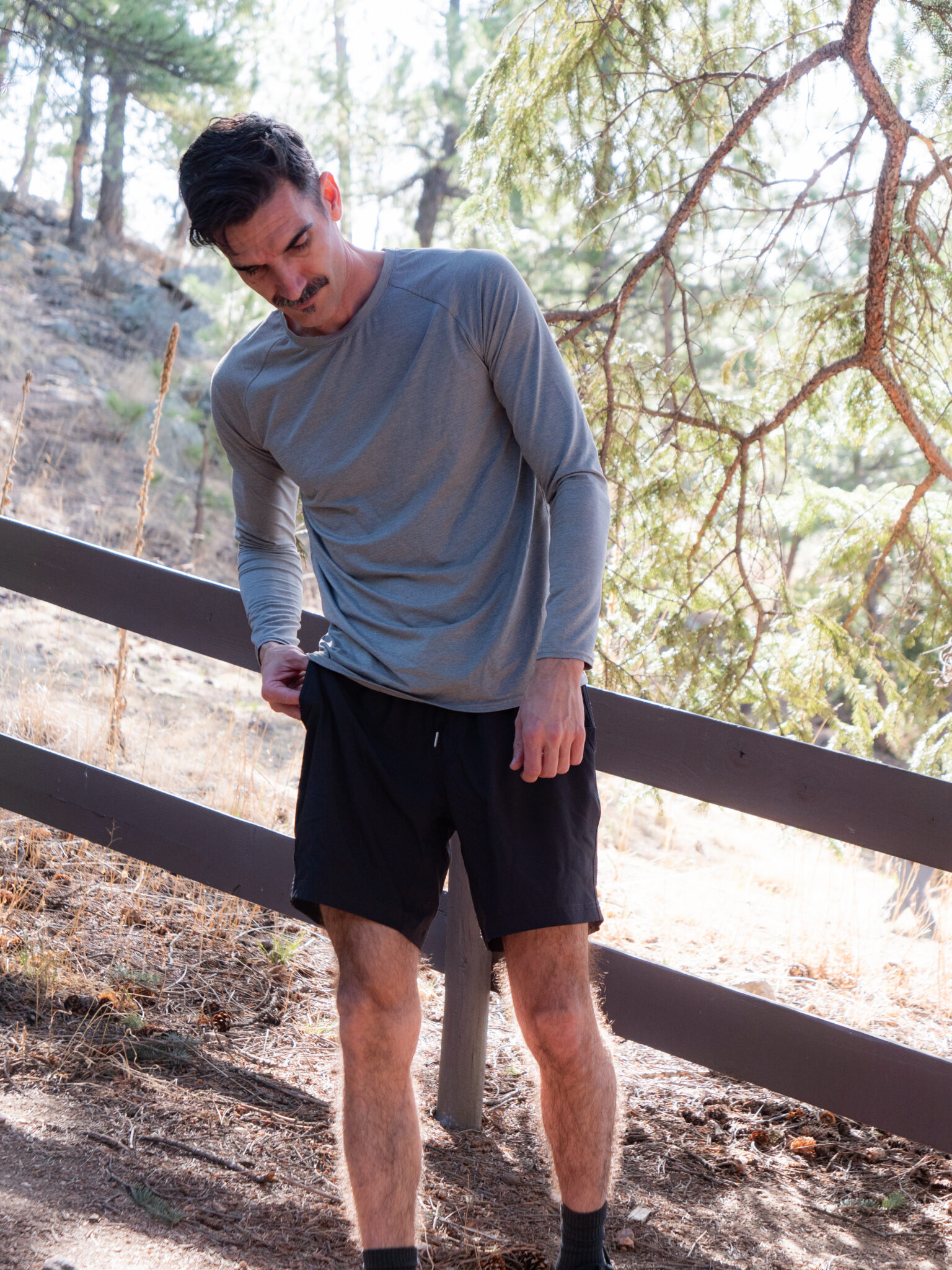 A man in a gray long-sleeve shirt and black shorts stands outdoors on a dirt path near a wooden fence and pine trees, looking down and adjusting his shirt.