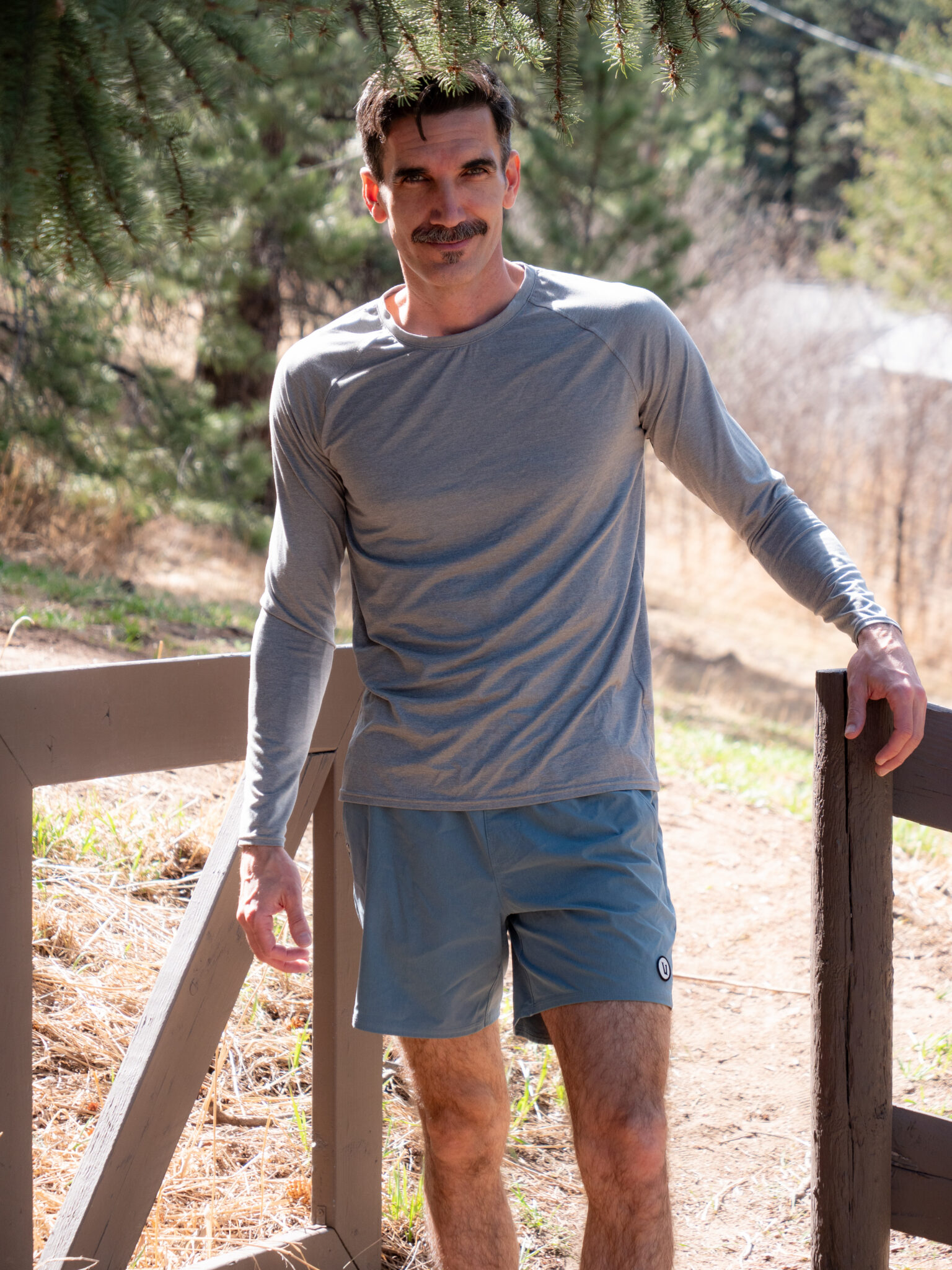 A man in a gray long-sleeve shirt and blue shorts stands outdoors near a wooden railing, with trees and dry grass in the background.