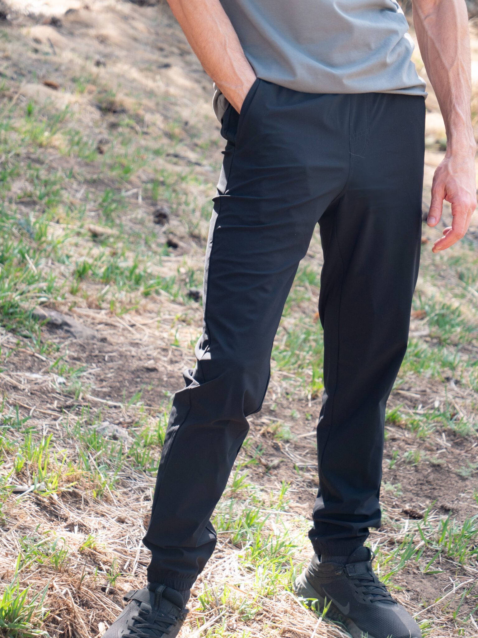 A person standing outdoors with hands in the pockets of black jogger pants and wearing a gray shirt and black shoes.