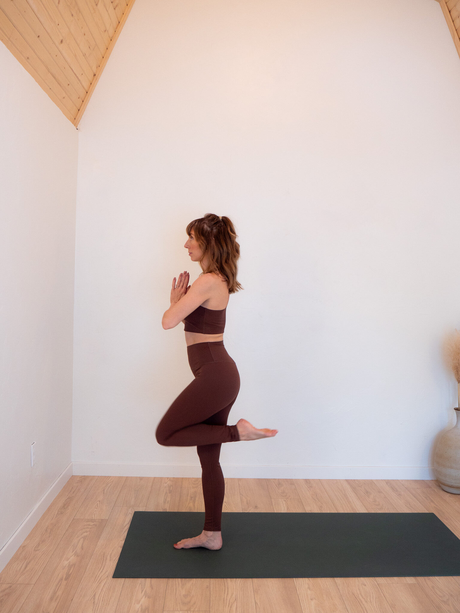 A woman practices yoga indoors, standing on one leg in a balancing pose on a yoga mat, with hands together at her chest.