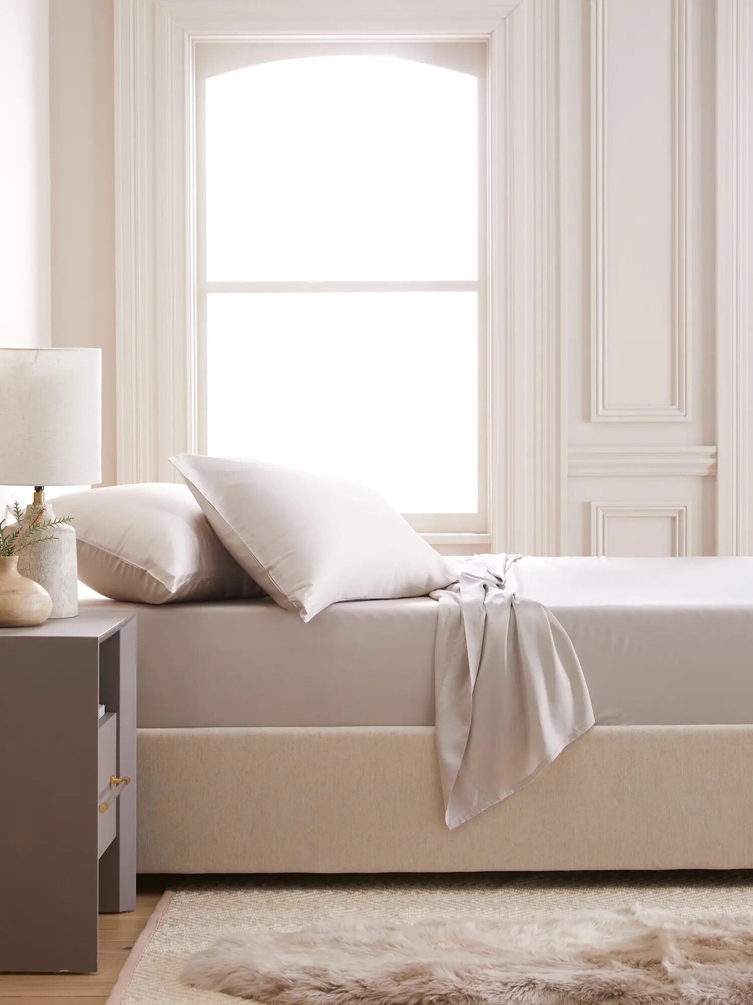 A neatly made bed with light-colored bedding is next to a nightstand with a lamp in a bright room featuring large windows and elegant trim.
