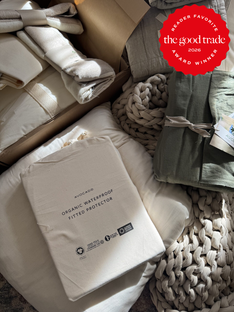 A collection of bedding items including an Avocado organic waterproof fitted protector, folded blankets, and sheets, displayed in natural light. A "Good Trade 2026 Award Winner" badge is shown.