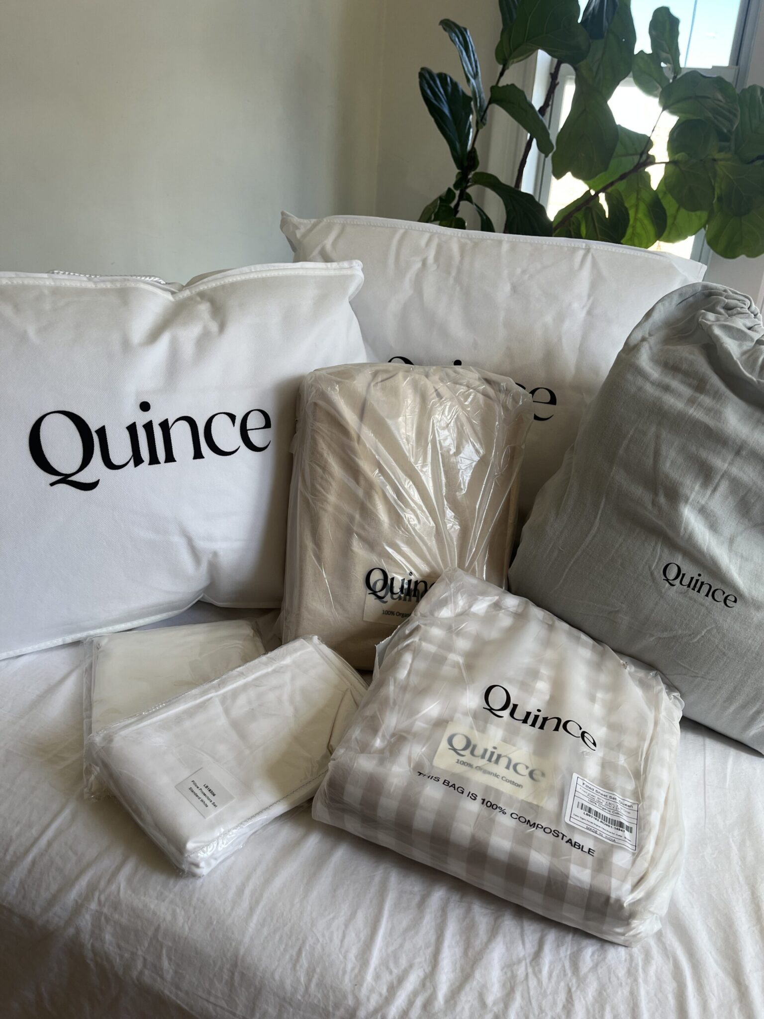 A collection of Quince bedding items, including pillows, sheets, and pillowcases, arranged on a bed near a window with a plant in the background.