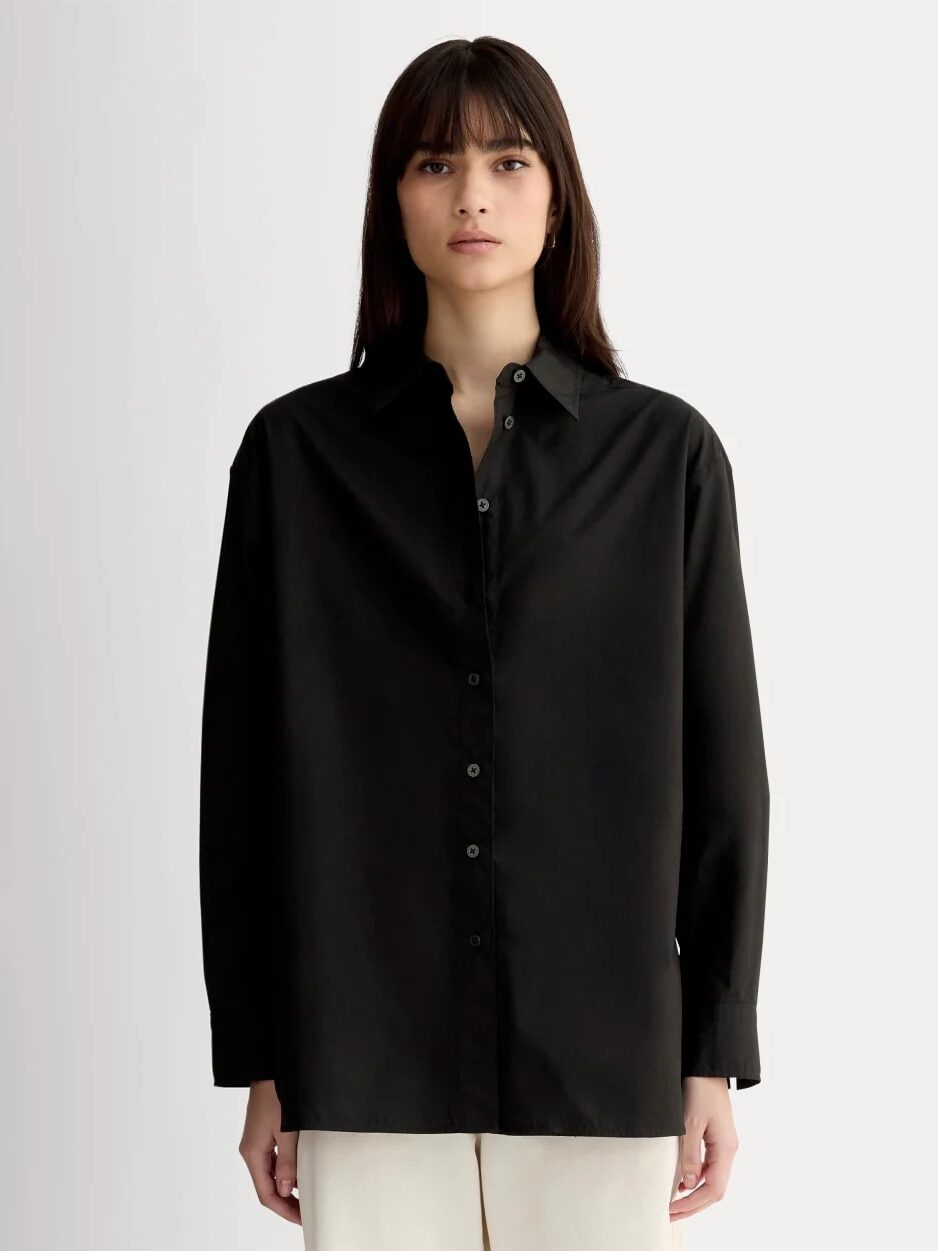 A person with straight dark hair wears a loose-fitting black button-up shirt and light-colored pants, standing against a plain light background.