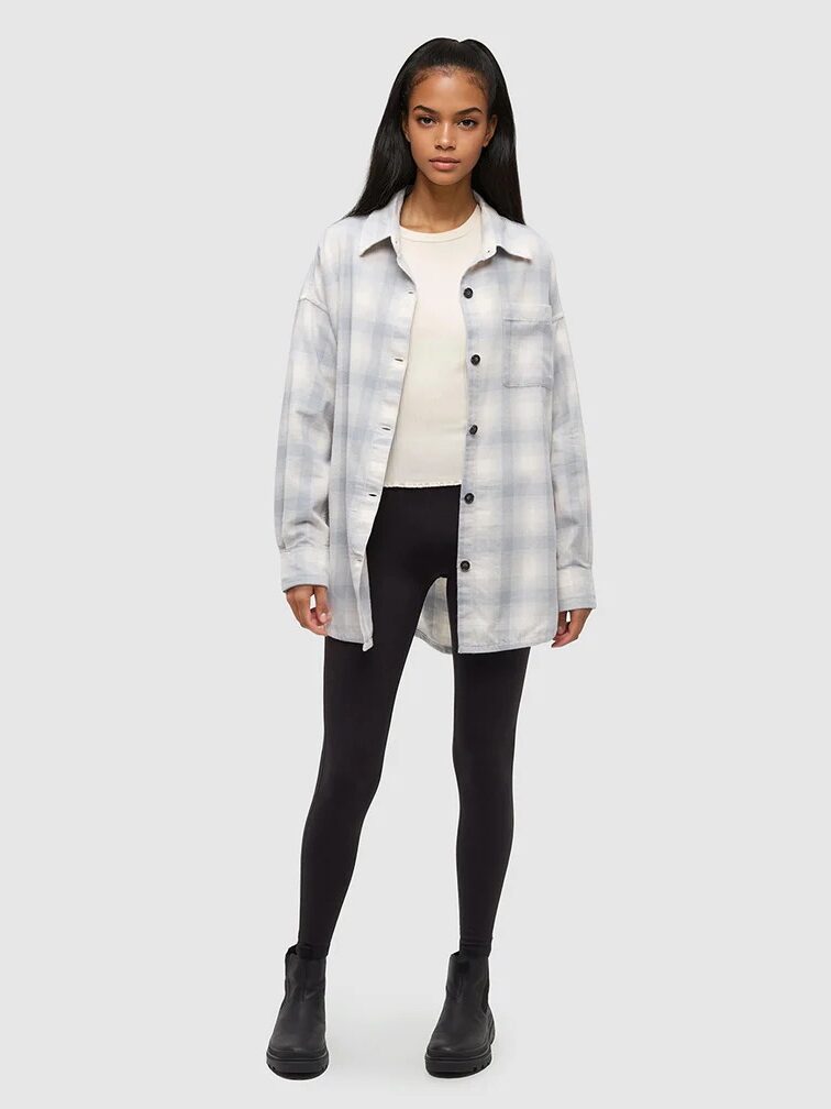 A person stands facing forward wearing a light plaid button-up shirt over a white top, black leggings, and black ankle boots against a plain light background.