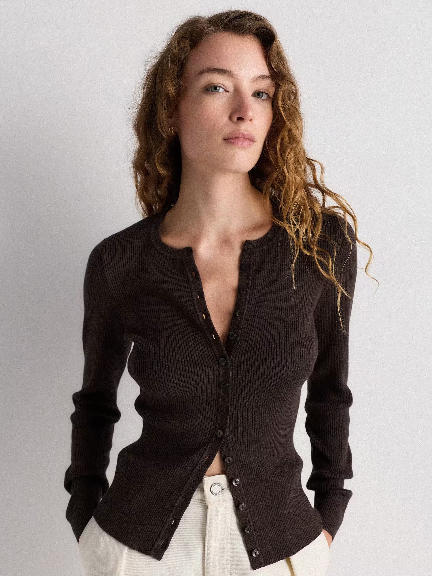 A woman with wavy hair wearing a dark ribbed button-up cardigan and light-colored pants stands against a plain background.
