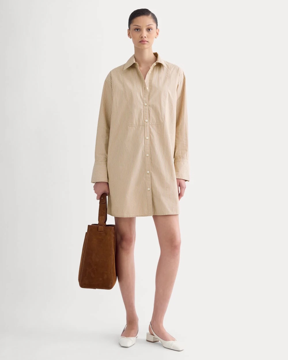 A woman stands facing forward wearing a beige striped shirt dress, white flats, and holding a brown suede handbag against a plain white background.
