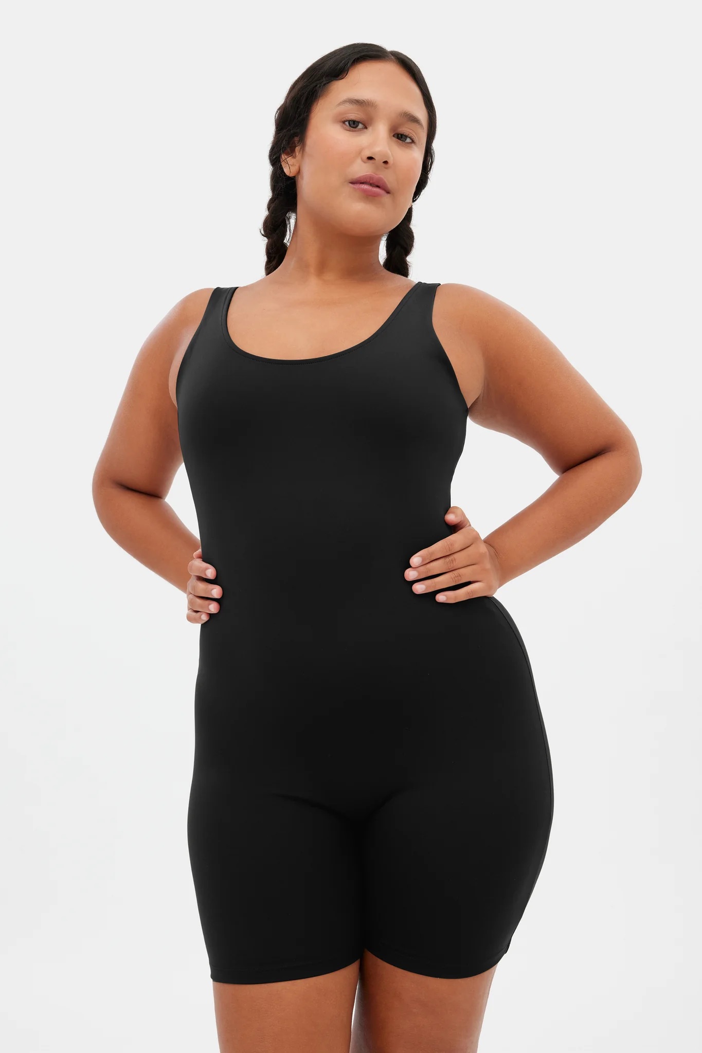 A woman stands with hands on hips, wearing a black sleeveless one-piece bodysuit against a plain white background.