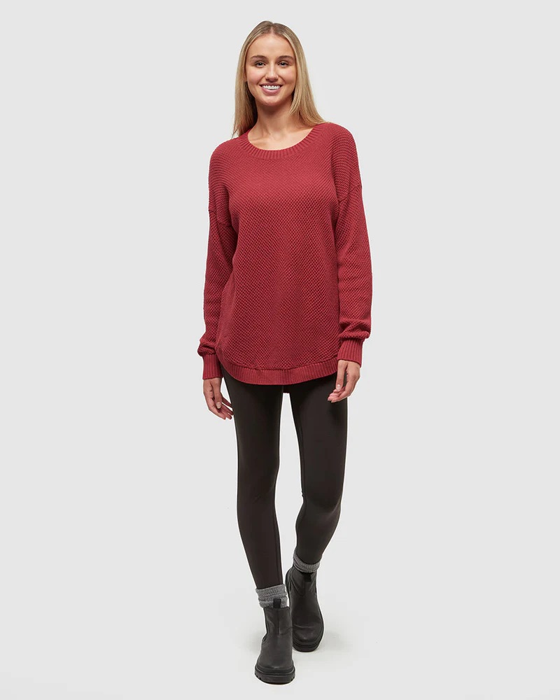 A woman stands against a plain background wearing a textured red sweater, black leggings, and black ankle boots with gray socks. She is smiling and facing forward.