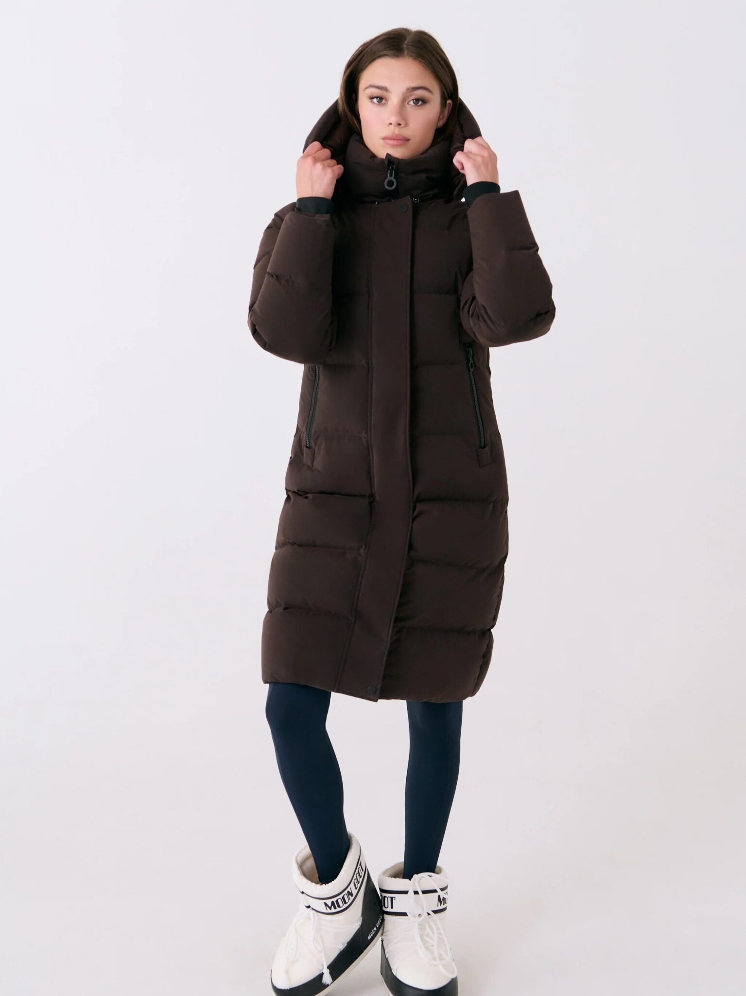 A woman wears a long, dark brown puffer coat with a hood, dark leggings, and white winter boots, standing against a plain light background.
