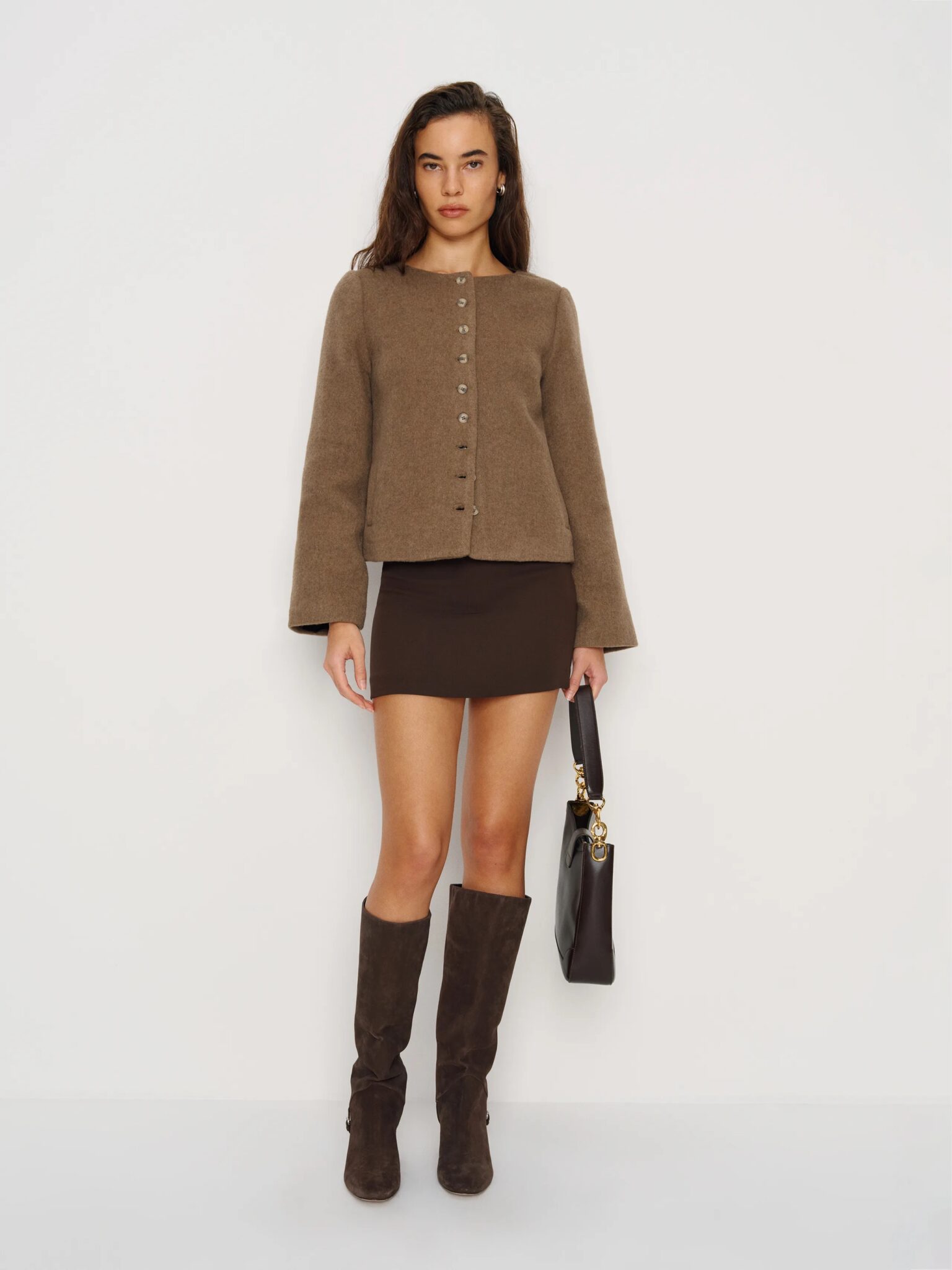 A woman stands against a white background wearing a brown buttoned jacket, brown mini skirt, brown knee-high boots, and holding a black handbag.