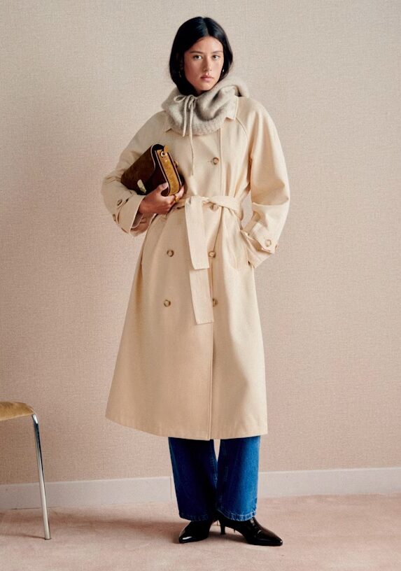 A woman wearing a beige trench coat over a gray hoodie, blue jeans, and black shoes, holding a brown clutch, stands indoors next to a chair.