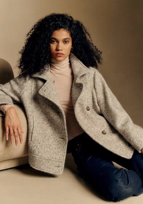 A person with long curly hair wears a light gray double-breasted coat over a beige turtleneck, sitting on the floor next to a tan sofa, with a neutral background.