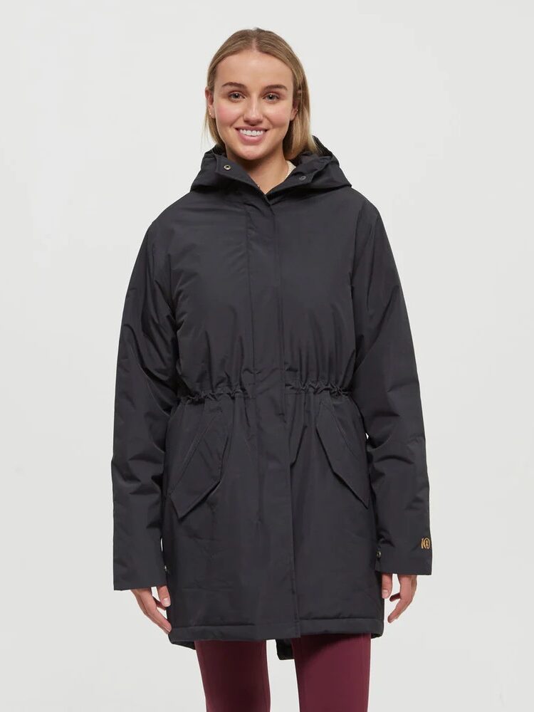 A woman stands facing forward, wearing a black hooded parka with front pockets and burgundy leggings against a plain light background.