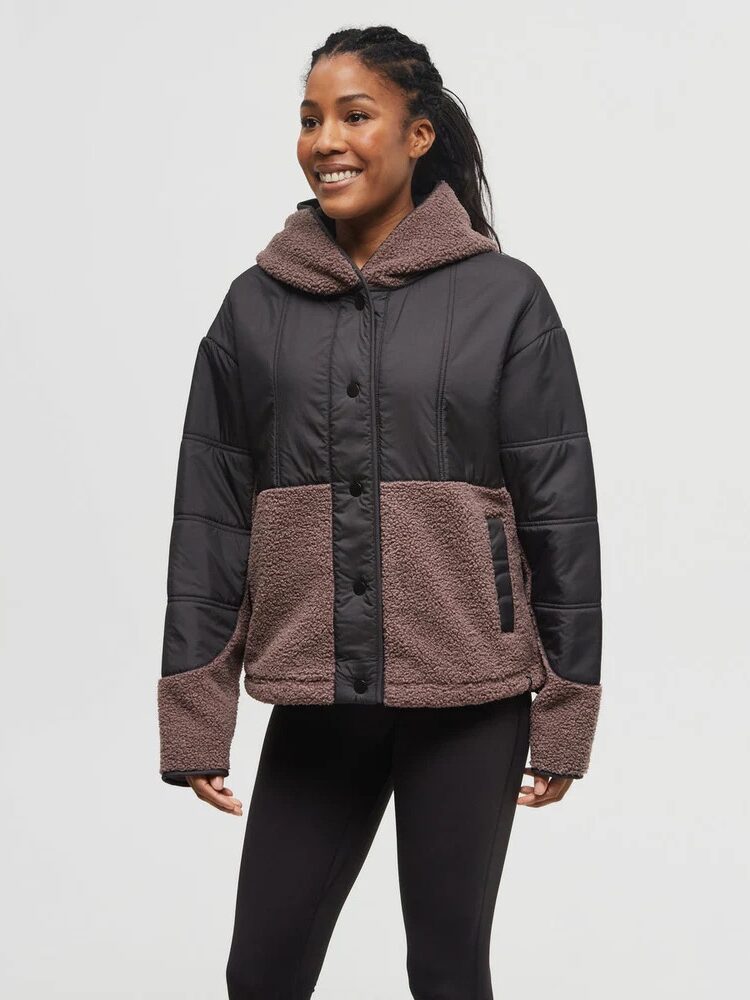 A woman wearing a black and brown hooded jacket with a button front and side pockets, paired with black leggings, stands against a plain light background.