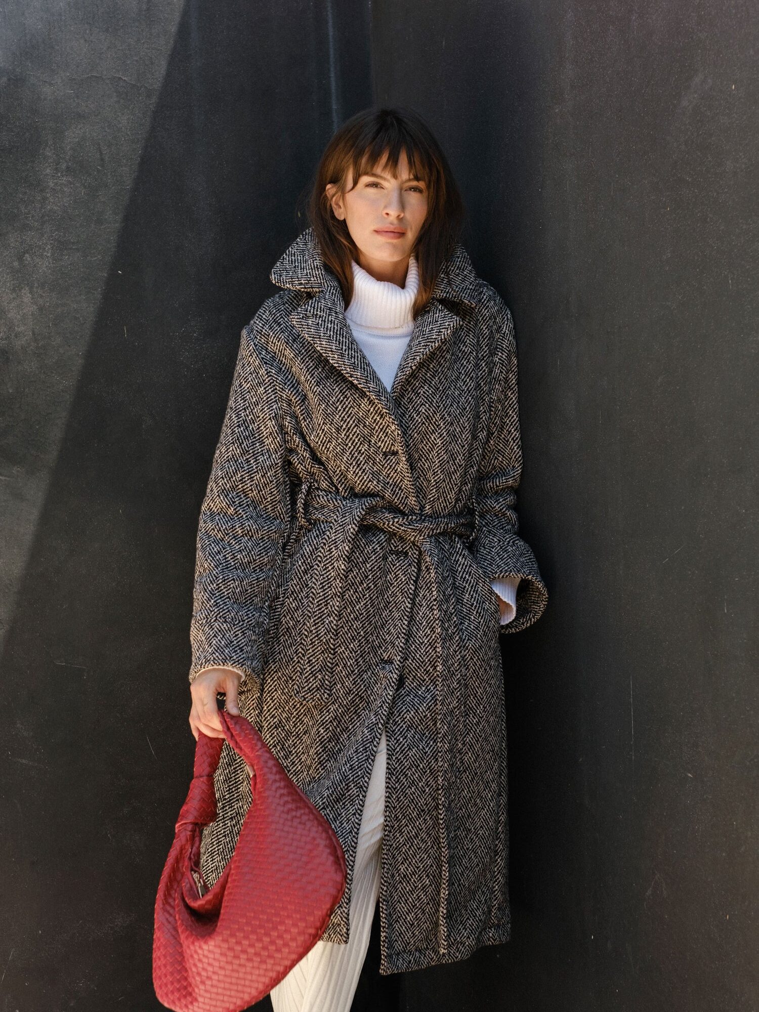 Woman in a gray belted coat and white turtleneck stands against a dark wall, holding a textured red handbag in her left hand.